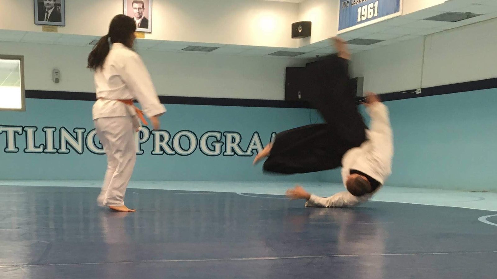 A student in hakama flies through a breakfall (ukemi) across the blue mat, feet in the air and one hand reaching for the ground, while their training partner stands where the throw ended