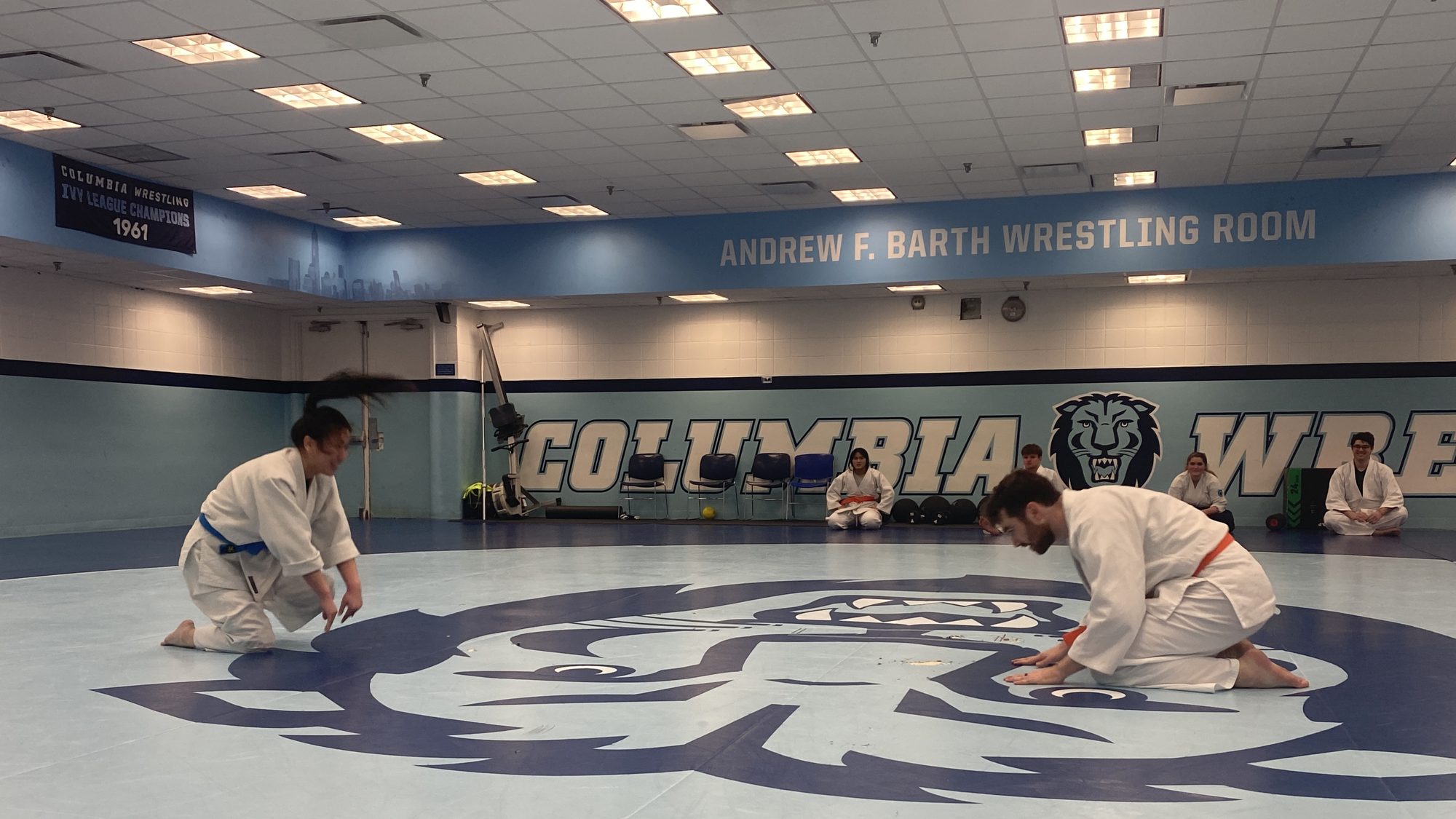 Two members in white gi kneel facing each other on the Columbia lion at the center of the wrestling-room mat, hands to the floor in a seated bow, with a few classmates seated along the wall behind